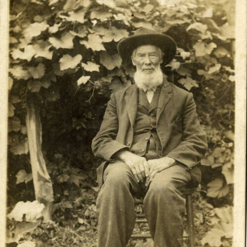 Historic photograph of a man sitting in a chair outside in front of a bush, wearing a dark suit and hat with a white beard.